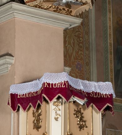 Zdjęcie nr 2: Ambona przyścienna wisząca, z dostępem od strony prezbiterium w postaci dwubiegowych schodów z podestem. Balustrada pełna, na cokole, dekorowana płycinami w formie rombów, z poręczą w postaci gzymsu. Kosz na planie kwadratu o ściętych narożach i wklęsłych bokach, na cokole w formie spłaszczonego wałka dekorowanego kampanulami w układzie poziomym. U spodu podwieszony odwrócony ostrosłup o ściętym wierzchołku i wklęsłych bokach ujęty w narożach spływami wolutowymi, dekorowany kampanulami, zakończony szyszką zdobioną cęgami. Ścianki kosza wydzielone pilastrami w formie wolut, których trzony dekorowane są ukwieconymi gałązkami. Na ściankach rocaille'owe płyciny z ornamentem roślinnym. Parapet w postaci gzymsu. Baldachim na planie analogicznym względem kosza, w formie gzymsu, dekorowany stylizowanym perełkowaniem, z podwieszonym lambrekinem. Na nim, na podstawie utworzonej z obłoków figura Chrystusa, zwróconego frontalnie, trzymającego na lewym ramieniu krzyż, z prawą ręką wskazującą w górę. Po jego lewej figurka Mojżesza z tablicami przykazań, po prawej św. Michała Archanioła trzymającego trąbę i wagę. Na podniebieniu baldachimu płaskorzeźbiona gołębica Ducha Świętego z czterema wiązkami promieni oraz dwie ukwiecone gałązki. Struktura polichromowana w kolorze beżowym, z białym detalem, ornamentyka i profile złocone, obłoki i gołębica srebrzone.