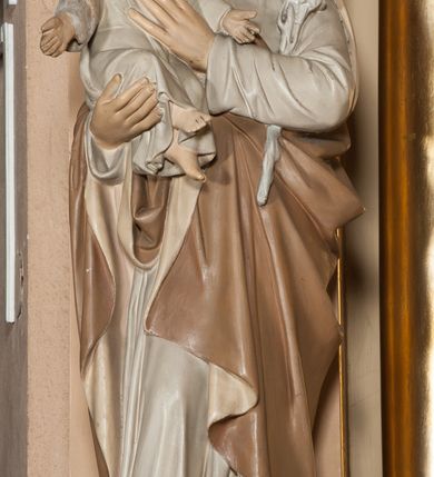 Zdjęcie nr 1: Rzeźba pełnoplastyczna, ustawiona na profilowanej podstawie na planie kwadratu o ściętych narożach. Józef ustawiony jest frontalnie, w kontrapoście, z lekko pochyloną głową. Na prawym przedramieniu podtrzymuje siedzące Dzieciątko, lewym przedramieniem przyciska do ciała gałązkę lilii, kładąc dłoń na piersi Dzieciątka. Twarz świętego jest pociągła, z wysokim czołem, o delikatnych rysach, małych oczach skierowanych w dół i prostym nosie, okolona krótką brodą i krótkimi kręconymi włosami. Ubrany jest w białą tunikę oraz jasnobrązowy płaszcz z beżową podszewką zarzucony na prawe ramię i otaczający postać. Dzieciątko skierowane jest w trzech czwartych w lewo, ma pochyloną głowę i rozłożone na boki ręce. Twarz ma okrągłą o pełnych policzkach i delikatnych rysach, okoloną krótkimi, kręconymi włosami. Ubrane jest w białą, luźną tunikę. Polichromia ciała naturalistyczna. 