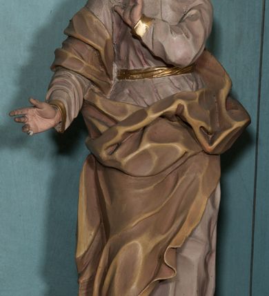 Zdjęcie nr 6: Rzeźba drążona, ustawiona na niewielkiej podstawie na planie kwadratu. Figura ukazana frontalnie, w kontrapoście, z głową przechyloną na prawy bark. Lewą rękę ma ugiętą przy piersi, prawą wyciąga w bok. Twarz ma pociągłą, o starczych rysach, dużych oczach, lekko garbatym nosie i niewielkich ustach, okoloną długimi włosami, zakrytymi welonem. Ubrana jest w bladoróżową tunikę o pozłoconych brzegach rękawów, przepasaną pozłoconą szarfą, silnie drapowaną oraz jasnobrązowy płaszcz zawinięty na prawym ramieniu i otaczający postać w partii bioder, o ekspresyjnie kształtowanej tkaninie. Na głowie ma jasnobrązowy welon opadający na plecy. Polichromia ciała naturalistyczna. 
