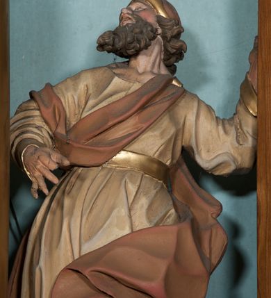 Zdjęcie nr 7: Rzeźba drążona, ustawiona na niewysokiej podstawie o nieregularnym kształcie. Figura zwrócona w trzech czwartych w prawo, w kontrapoście, sylwetka silnie odchylona w tył, z głową skierowaną do góry. Prawa ręka ugięta, znajduje się przed klatką piersiową, lewa wyciągnięta jest w lewo i w górę. Twarz ma pociągłą, o skierowanych w górę oczach, lekko garbatym nosie i otwartych ustach, okolona jest wysuniętą w przód, gęstą, kręconą brodą oraz krótkimi włosami rzeźbionymi w pojedyncze pukle. Święty ubrany jest w beżową tunikę o pozłoconych mankietach i pasie w talii oraz pozłacane nakrycie głowy. Przez klatkę piersiową i prawe ramię przerzucony jest fragment czerwono-brązowego, silnie drapowanego płaszcza, który opada na plecy i lewą nogę. Polichromia ciała naturalistyczna. 