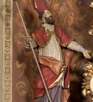 Zdjęcie nr 5: Rzeźba drążona, ustawiona na podstawie na planie prostokąta. Figura zwrócona jest w trzech czwartych w lewo, w delikatnym kontrapoście, o łukowato wygiętej sylwetce. W prawej, ugiętej ręce trzyma pastorał, lewą wyciąga przed siebie. Twarz ma pociągłą, szczupłą, z silnie zaznaczonymi łukami brwiowymi i kośćmi policzkowymi, z szeroko otwartymi oczami, prostym nosem i otwartymi ustami, okoloną bujną, kręconą brodą oraz krótkimi, kręconymi włosami. Święty jest ubrany w fioletową albę, białą rokietę ze złotą lamówką u szyi oraz czerwoną kapę ze złotą lamówką i pomarańczową podszewką, zapiętą na piersi, opadającą na plecy, z zawiniętym i ekspresyjnie rozwianym fragmentem tkaniny na prawej nodze. Na głowie wysoka infuła z pozłoconym krzyżem i brzegami. Polichromia naturalistyczna w partiach ciała. 