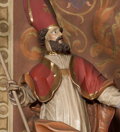 Zdjęcie nr 4: Rzeźba drążona, ustawiona na podstawie na planie prostokąta. Figura zwrócona jest w trzech czwartych w lewo, w delikatnym kontrapoście, o łukowato wygiętej sylwetce. W prawej, ugiętej ręce trzyma pastorał, lewą wyciąga przed siebie. Twarz ma pociągłą, szczupłą, z silnie zaznaczonymi łukami brwiowymi i kośćmi policzkowymi, z szeroko otwartymi oczami, prostym nosem i otwartymi ustami, okoloną bujną, kręconą brodą oraz krótkimi, kręconymi włosami. Święty jest ubrany w fioletową albę, białą rokietę ze złotą lamówką u szyi oraz czerwoną kapę ze złotą lamówką i pomarańczową podszewką, zapiętą na piersi, opadającą na plecy, z zawiniętym i ekspresyjnie rozwianym fragmentem tkaniny na prawej nodze. Na głowie wysoka infuła z pozłoconym krzyżem i brzegami. Polichromia naturalistyczna w partiach ciała. 