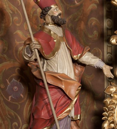 Zdjęcie nr 6: Rzeźba drążona, ustawiona na podstawie na planie prostokąta. Figura zwrócona jest w trzech czwartych w lewo, w delikatnym kontrapoście, o łukowato wygiętej sylwetce. W prawej, ugiętej ręce trzyma pastorał, lewą wyciąga przed siebie. Twarz ma pociągłą, szczupłą, z silnie zaznaczonymi łukami brwiowymi i kośćmi policzkowymi, z szeroko otwartymi oczami, prostym nosem i otwartymi ustami, okoloną bujną, kręconą brodą oraz krótkimi, kręconymi włosami. Święty jest ubrany w fioletową albę, białą rokietę ze złotą lamówką u szyi oraz czerwoną kapę ze złotą lamówką i pomarańczową podszewką, zapiętą na piersi, opadającą na plecy, z zawiniętym i ekspresyjnie rozwianym fragmentem tkaniny na prawej nodze. Na głowie wysoka infuła z pozłoconym krzyżem i brzegami. Polichromia naturalistyczna w partiach ciała. 