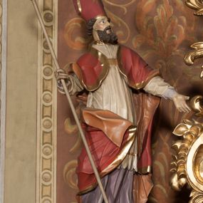 Zdjęcie nr 1: Rzeźba drążona, ustawiona na podstawie na planie prostokąta. Figura zwrócona jest w trzech czwartych w lewo, w delikatnym kontrapoście, o łukowato wygiętej sylwetce. W prawej, ugiętej ręce trzyma pastorał, lewą wyciąga przed siebie. Twarz ma pociągłą, szczupłą, z silnie zaznaczonymi łukami brwiowymi i kośćmi policzkowymi, z szeroko otwartymi oczami, prostym nosem i otwartymi ustami, okoloną bujną, kręconą brodą oraz krótkimi, kręconymi włosami. Święty jest ubrany w fioletową albę, białą rokietę ze złotą lamówką u szyi oraz czerwoną kapę ze złotą lamówką i pomarańczową podszewką, zapiętą na piersi, opadającą na plecy, z zawiniętym i ekspresyjnie rozwianym fragmentem tkaniny na prawej nodze. Na głowie wysoka infuła z pozłoconym krzyżem i brzegami. Polichromia naturalistyczna w partiach ciała. 