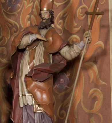 Zdjęcie nr 3: Rzeźba drążona, ustawiona na niskiej podstawie na planie prostokąta. Figura zwrócona jest w trzech czwartych w prawo, w delikatnym kontrapoście, o łukowatym układzie sylwetki, odchylona w tył. Prawa ręka skierowana na pierś, w lewej, ugiętej ferula. Twarz ma pociągłą, szczupłą, z wyraźnie zaznaczonymi kośćmi policzkowymi i łukami brwiowymi, z głęboko osadzonymi oczami, prostym nosem i otwartymi ustami, okoloną długą, falowaną i gęstą brodą. Włosy krótkie, kręcone, opadające na tył głowy. Święty ubrany jest w fioletową albę, białą rokietę, długą pomarańczową stułę z pozłacanymi brzegami oraz pomarańczową kapę z ciemnoróżową podszewką, zapiętą na piersi, opadającą na plecy, z rozwianym i zawiniętym na lewej nodze fragmentem tkaniny. Na głowie wysoka pomarańczowa infuła z pozłacanym krzyżem i brzegami. Polichromia ciała naturalistyczna. 