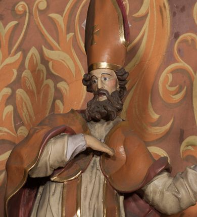 Zdjęcie nr 5: Rzeźba drążona, ustawiona na niskiej podstawie na planie prostokąta. Figura zwrócona jest w trzech czwartych w prawo, w delikatnym kontrapoście, o łukowatym układzie sylwetki, odchylona w tył. Prawa ręka skierowana na pierś, w lewej, ugiętej ferula. Twarz ma pociągłą, szczupłą, z wyraźnie zaznaczonymi kośćmi policzkowymi i łukami brwiowymi, z głęboko osadzonymi oczami, prostym nosem i otwartymi ustami, okoloną długą, falowaną i gęstą brodą. Włosy krótkie, kręcone, opadające na tył głowy. Święty ubrany jest w fioletową albę, białą rokietę, długą pomarańczową stułę z pozłacanymi brzegami oraz pomarańczową kapę z ciemnoróżową podszewką, zapiętą na piersi, opadającą na plecy, z rozwianym i zawiniętym na lewej nodze fragmentem tkaniny. Na głowie wysoka pomarańczowa infuła z pozłacanym krzyżem i brzegami. Polichromia ciała naturalistyczna. 