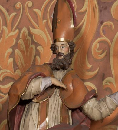 Zdjęcie nr 4: Rzeźba drążona, ustawiona na niskiej podstawie na planie prostokąta. Figura zwrócona jest w trzech czwartych w prawo, w delikatnym kontrapoście, o łukowatym układzie sylwetki, odchylona w tył. Prawa ręka skierowana na pierś, w lewej, ugiętej ferula. Twarz ma pociągłą, szczupłą, z wyraźnie zaznaczonymi kośćmi policzkowymi i łukami brwiowymi, z głęboko osadzonymi oczami, prostym nosem i otwartymi ustami, okoloną długą, falowaną i gęstą brodą. Włosy krótkie, kręcone, opadające na tył głowy. Święty ubrany jest w fioletową albę, białą rokietę, długą pomarańczową stułę z pozłacanymi brzegami oraz pomarańczową kapę z ciemnoróżową podszewką, zapiętą na piersi, opadającą na plecy, z rozwianym i zawiniętym na lewej nodze fragmentem tkaniny. Na głowie wysoka pomarańczowa infuła z pozłacanym krzyżem i brzegami. Polichromia ciała naturalistyczna. 