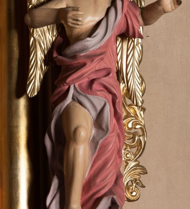 Zdjęcie nr 1: Rzeźba ścięta z tyłu, drążona, przedstawia anioła stojącego na  rzeźbionym, posrebrzanym obłoku. Figura zwrócona frontalnie, z prawą nogą opartą na obłoku, głową przechyloną na prawy bark oraz uniesionymi przed sobą, ugiętymi rękami. Twarz owalna, o pełnych policzkach i wystającej brodzie, migdałowatych, opadających oczach, małym nosie i ustach, okolona krótkimi, zaczesanymi do tyłu włosami. Anioł ubrany jest w silnie fałdowaną, różową tunikę z fioletową podszewką, z jednym rękawem na lewym przedramieniu, odsłaniającą połowę torsu i prawe ramię oraz prawą nogę. Skrzydła pozłocone, złożone po bokach postaci. Polichromia ciała naturalistyczna. 

