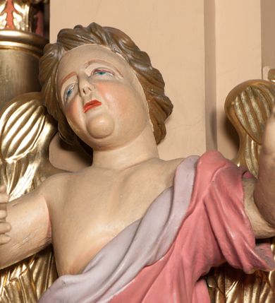 Zdjęcie nr 2: Rzeźba ścięta z tyłu, drążona, przedstawia anioła stojącego na  rzeźbionym, posrebrzanym obłoku. Figura zwrócona frontalnie, z prawą nogą opartą na obłoku, głową przechyloną na prawy bark oraz uniesionymi przed sobą, ugiętymi rękami. Twarz owalna, o pełnych policzkach i wystającej brodzie, migdałowatych, opadających oczach, małym nosie i ustach, okolona krótkimi, zaczesanymi do tyłu włosami. Anioł ubrany jest w silnie fałdowaną, różową tunikę z fioletową podszewką, z jednym rękawem na lewym przedramieniu, odsłaniającą połowę torsu i prawe ramię oraz prawą nogę. Skrzydła pozłocone, złożone po bokach postaci. Polichromia ciała naturalistyczna. 

