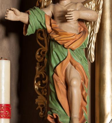 Zdjęcie nr 1: Rzeźba ścięta z tyłu, drążona, ustawiona na rzeźbionym, posrebrzanym obłoku. Figura anioła ukazana frontalnie, z lewą nogą opartą na obłoku oraz wyciągniętymi przed siebie, ugiętymi rękoma. Twarz ma owalną, o pełnych policzkach i podkreślonej brodzie, migdałowatych, opadających oczach, małym nosie i ustach, okoloną krótkimi, falowanymi, zaczesanymi w tył włosami. Anioł ubrany jest w obficie fałdowaną zieloną tunikę z pomarańczową podszewką, z jednym rękawem na prawym ramieniu, odsłaniającą tors, lewe ramię oraz lewą nogę. Skrzydła pozłacane, złożone po bokach postaci. Polichromia ciała naturalistyczna. 

