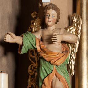 Zdjęcie nr 1: Rzeźba ścięta z tyłu, drążona, ustawiona na rzeźbionym, posrebrzanym obłoku. Figura anioła ukazana frontalnie, z lewą nogą opartą na obłoku oraz wyciągniętymi przed siebie, ugiętymi rękoma. Twarz ma owalną, o pełnych policzkach i podkreślonej brodzie, migdałowatych, opadających oczach, małym nosie i ustach, okoloną krótkimi, falowanymi, zaczesanymi w tył włosami. Anioł ubrany jest w obficie fałdowaną zieloną tunikę z pomarańczową podszewką, z jednym rękawem na prawym ramieniu, odsłaniającą tors, lewe ramię oraz lewą nogę. Skrzydła pozłacane, złożone po bokach postaci. Polichromia ciała naturalistyczna. 
