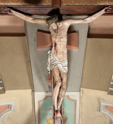 Zdjęcie nr 2: Figura umarłego Chrystusa przybita do krzyża o prostych ramionach trzema gwoździami. Ciało w lekkim zwisie o horyzontalnie rozłożonych ramionach, głowie opadającej bezwładnie w prawą stronę, z lekko ugiętymi w kolanach nogami oraz stopami skrzyżowanymi w układzie prawa na lewą. Twarz szczupła, pociągła, z zamkniętymi oczami, długim, wąskim nosem i otwartymi ustami, okolona krótką brodą skręconą w regularne pukle oraz falowanymi włosami opadającymi na plecy i prawe ramię. Na głowie ma zieloną koronę cierniową w formie kołpaka. Sylwetka smukła, proporcjonalnie wydłużona, ciało silnie wychudzone o podkreślonych mięśniach, żebrach i mostku klatki piersiowej oraz ścięgnach nóg i rąk. W boku zaznaczona rana z rzeźbionymi kroplami krwi. Perizonium   białe, krótkie, ściśle przylegające do ciała, silnie drapowane, odwinięte i osuwające się w partii podbrzusza, z kaskadowym zwisem tkaniny na prawym boku. Polichromia ciała naturalistyczna z zaznaczonymi śladami męki. Do górnej belki krzyża przybita płaskorzeźbiona tabliczka z inskrypcją „INRI”. 