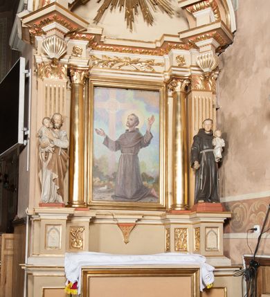 Zdjęcie nr 3: Obraz w formie stojącego owalu przedstawia św. Antoniego Padewskiego, któremu wśród obłoków ukazała się Matka Boska, przekazując mu w ramiona Dzieciątko. W prawej części kompozycji święty w pozycji klęczącej, skierowany w trzech czwartych w prawo, z pochyloną głową oraz wyciągniętymi przed siebie ramionami, w których trzyma Dzieciątko. Ubrany jest w brązowy habit franciszkański z peleryną i kapturem, przewiązany w pasie sznurem, na głowie ma tonsurę. Dzieciątko zwrócone w trzech czwartych w lewo, z wyciągniętymi w stronę świętego rękami, owinięte w pieluszki. Przed Antonim stolik nakryty żółtą tkaniną z leżącą księgą oraz gałązką lilii. Nad nim znajduje się Matka Boska siedząca na obłokach, zwrócona w trzech czwartych w lewo, pochylona, z wyciągniętymi w stronę świętego rękami. Ubrana jest w czerwoną suknię przepasaną w talii oraz niebieski płaszcz zawinięty na lewym ramieniu, rozwiany przy prawym boku. W dolnej części obrazu widoczna bladoróżowa podłoga, w górnej rozświetlone, żółte niebo, po prawej stronie obrazu na obłokach dwie uskrzydlone główki anielskie. 