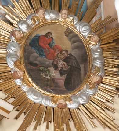 Zdjęcie nr 2: Obraz w formie stojącego owalu przedstawia św. Antoniego Padewskiego, któremu wśród obłoków ukazała się Matka Boska, przekazując mu w ramiona Dzieciątko. W prawej części kompozycji święty w pozycji klęczącej, skierowany w trzech czwartych w prawo, z pochyloną głową oraz wyciągniętymi przed siebie ramionami, w których trzyma Dzieciątko. Ubrany jest w brązowy habit franciszkański z peleryną i kapturem, przewiązany w pasie sznurem, na głowie ma tonsurę. Dzieciątko zwrócone w trzech czwartych w lewo, z wyciągniętymi w stronę świętego rękami, owinięte w pieluszki. Przed Antonim stolik nakryty żółtą tkaniną z leżącą księgą oraz gałązką lilii. Nad nim znajduje się Matka Boska siedząca na obłokach, zwrócona w trzech czwartych w lewo, pochylona, z wyciągniętymi w stronę świętego rękami. Ubrana jest w czerwoną suknię przepasaną w talii oraz niebieski płaszcz zawinięty na lewym ramieniu, rozwiany przy prawym boku. W dolnej części obrazu widoczna bladoróżowa podłoga, w górnej rozświetlone, żółte niebo, po prawej stronie obrazu na obłokach dwie uskrzydlone główki anielskie. 