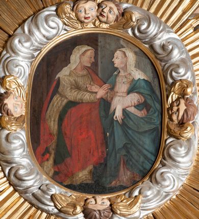 Zdjęcie nr 2: Obraz w formie stojącego owalu. Przedstawia spotkanie Marii i św. Elżbiety. W prawej części obrazu Maria zwrócona w trzech czwartych w prawo, lewą ręką podtrzymuje płaszcz na wysokości łona, prawą kieruje w stronę Elżbiety. Twarz ma owalną o bladej karnacji, łagodnych, młodzieńczych rysach i jasnych włosach ukrytych pod welonem. Ubrana jest w jasnoróżową suknię przepasaną w talii, niebieski płaszcz zarzucony na ramiona i plecy, podtrzymywany z przodu oraz biały welon opadający na ramiona i plecy. Nad głową ma nimb. W lewej części obrazu św. Elżbieta zwrócona w trzech czwartych w lewo, prawą rękę wyciąga w stronę Marii, lewą ją obejmuje. Twarz ma owalną o łagodnych, młodzieńczych rysach, jasnej karnacji, okoloną jasnymi włosami, przesłoniętymi welonem. Ubrana jest w beżową suknię, przepasaną w talii, czerwony płaszcz z zieloną podszewką, zarzucony na lewe ramię i opadający na plecy i z przodu postaci, na głowie ma biały welon. Postacie ukazane na zewnątrz przed wejściem do domu widocznego za Elżbietą. 
