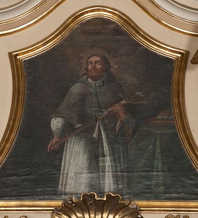 Zdjęcie nr 2: Obraz w formie trapezu o wklęsłych bokach, zamknięty faliście. W centrum kompozycji św. Jan Nepomucen ukazany do wysokości kolan, zwrócony frontalnie, z głową uniesioną do góry. Obiema rękami przytrzymuje krucyfiks oparty na jego lewym ramieniu. Twarz ma pociągłą, szczupłą, o rysach wydobytych głębokim światłocieniem, z oczami skierowanymi do góry, prostym nosem i pełnymi ustami, okoloną bujną brodą oraz włosami sięgającymi ramion. Wokół głowy świętego nimb i wieniec z pięciu gwiazd. Jan Nepomucen ubrany jest w białą komżę z wąskimi rękawami, lamowaną koronką, brązową almucję z różową podszewką zawiązaną u szyi niebieskim sznurem. W prawej części obrazu znajduje się stół przesłonięty zieloną tkaniną, z leżącymi na nim dwiema książkami. W górnej części dzieła ukazane ciemne niebo z zarysem obłoków, rozjaśnione wokół głowy świętego. Obraz ujęty profilowaną, pozłoconą ramą.