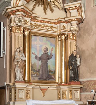 Zdjęcie nr 3: Nastawa architektoniczna, jednoosiowa, jednokondygnacyjna ze zwieńczeniem, ustawiona na wysokim dwustrefowym cokole. Ołtarz drewniany, prostopadłościenny z drewnianą mensą i kamiennym portatylem. Antepedium ujęte profilowaną ramą, w polu medalion z literą omega. Retabulum ujęte dwiema parami pilastrów, przed wewnętrzną stoi para kolumn o kapitelach korynckich. Zewnętrzne pilastry zdwojone, z kanelowanym trzonem, przed nimi znajdują się figury św. Józefa z Dzieciątkiem i św. Antoniego Padewskiego z Dzieciątkiem. Podpory dźwigają pełne belkowanie, dekorowane w partii fryzu ornamentem roślinnym. Nad figurami ustawione pod kątem imposty podtrzymywane przez wazonowate wsporniki, dźwigające przerwany półkolisty przyczółek. Całe pole środkowe wypełnia płycina w formie stojącego prostokąta z obrazem św. Franciszka, podtrzymywana u dołu przez wspornik. Zwieńczenie w formie trapezu wypełnione niemal w całości owalnym medalionem z obrazem św. Antoniego Padewskiego z Dzieciątkiem i Matką Boską, ujętym otokiem z obłoków z uskrzydlonymi główkami anielskimi w glorii promienistej. Struktura ołtarza polichromowana w kolorze beżowym, ornamentyka, profile i gloria pozłacane.