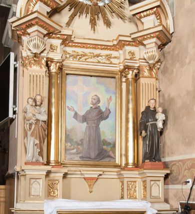 Zdjęcie nr 1: Nastawa architektoniczna, jednoosiowa, jednokondygnacyjna ze zwieńczeniem, ustawiona na wysokim dwustrefowym cokole. Ołtarz drewniany, prostopadłościenny z drewnianą mensą i kamiennym portatylem. Antepedium ujęte profilowaną ramą, w polu medalion z literą omega. Retabulum ujęte dwiema parami pilastrów, przed wewnętrzną stoi para kolumn o kapitelach korynckich. Zewnętrzne pilastry zdwojone, z kanelowanym trzonem, przed nimi znajdują się figury św. Józefa z Dzieciątkiem i św. Antoniego Padewskiego z Dzieciątkiem. Podpory dźwigają pełne belkowanie, dekorowane w partii fryzu ornamentem roślinnym. Nad figurami ustawione pod kątem imposty podtrzymywane przez wazonowate wsporniki, dźwigające przerwany półkolisty przyczółek. Całe pole środkowe wypełnia płycina w formie stojącego prostokąta z obrazem św. Franciszka, podtrzymywana u dołu przez wspornik. Zwieńczenie w formie trapezu wypełnione niemal w całości owalnym medalionem z obrazem św. Antoniego Padewskiego z Dzieciątkiem i Matką Boską, ujętym otokiem z obłoków z uskrzydlonymi główkami anielskimi w glorii promienistej. Struktura ołtarza polichromowana w kolorze beżowym, ornamentyka, profile i gloria pozłacane.