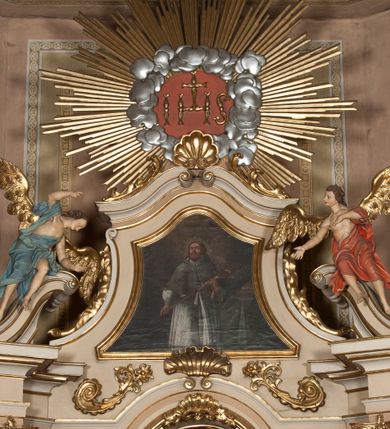 Zdjęcie nr 1: Rzeźby ścięte z tyłu, drążone, ukazane jako siedzące na przerwanym przyczółku, skierowane w stronę ołtarza. Lewa figura pochylona, z uniesioną nad głową prawą ręką i wyciągniętą przed siebie lewą. Figura anioła po prawej, lewą rękę kładzie na piersi, prawą wyciąga przed siebie. Anioły mają pociągłe twarze, z wystającymi brodami, dużymi oczami, zadartymi nosami i pełnymi ustami, okolone silnie kręconymi włosami opadającymi na kark. Anioł po lewej stronie ubrany jest w niebieską, silnie drapowaną szatę, odsłaniającą częściowo tors, przerzuconą przez prawe ramię, podtrzymywaną na lewym ramieniu wstążką, otaczającą postać w partii bioder. Anioł po prawej ubrany jest w czerwoną, ekspresyjnie fałdowaną szatę odsłaniającą tors, przerzuconą przez lewe ramię i otaczającą postać w partii bioder. Skrzydła obu aniołów rozpostarte na boki, pozłocone. Polichromia ciała naturalistyczna. 