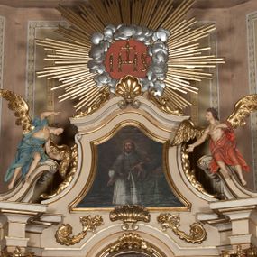 Zdjęcie nr 1: Rzeźby ścięte z tyłu, drążone, ukazane jako siedzące na przerwanym przyczółku, skierowane w stronę ołtarza. Lewa figura pochylona, z uniesioną nad głową prawą ręką i wyciągniętą przed siebie lewą. Figura anioła po prawej, lewą rękę kładzie na piersi, prawą wyciąga przed siebie. Anioły mają pociągłe twarze, z wystającymi brodami, dużymi oczami, zadartymi nosami i pełnymi ustami, okolone silnie kręconymi włosami opadającymi na kark. Anioł po lewej stronie ubrany jest w niebieską, silnie drapowaną szatę, odsłaniającą częściowo tors, przerzuconą przez prawe ramię, podtrzymywaną na lewym ramieniu wstążką, otaczającą postać w partii bioder. Anioł po prawej ubrany jest w czerwoną, ekspresyjnie fałdowaną szatę odsłaniającą tors, przerzuconą przez lewe ramię i otaczającą postać w partii bioder. Skrzydła obu aniołów rozpostarte na boki, pozłocone. Polichromia ciała naturalistyczna. 