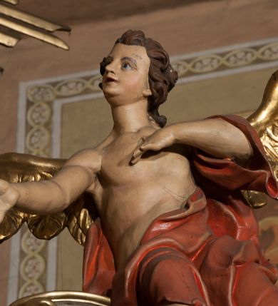 Zdjęcie nr 3: Rzeźby ścięte z tyłu, drążone, ukazane jako siedzące na przerwanym przyczółku, skierowane w stronę ołtarza. Lewa figura pochylona, z uniesioną nad głową prawą ręką i wyciągniętą przed siebie lewą. Figura anioła po prawej, lewą rękę kładzie na piersi, prawą wyciąga przed siebie. Anioły mają pociągłe twarze, z wystającymi brodami, dużymi oczami, zadartymi nosami i pełnymi ustami, okolone silnie kręconymi włosami opadającymi na kark. Anioł po lewej stronie ubrany jest w niebieską, silnie drapowaną szatę, odsłaniającą częściowo tors, przerzuconą przez prawe ramię, podtrzymywaną na lewym ramieniu wstążką, otaczającą postać w partii bioder. Anioł po prawej ubrany jest w czerwoną, ekspresyjnie fałdowaną szatę odsłaniającą tors, przerzuconą przez lewe ramię i otaczającą postać w partii bioder. Skrzydła obu aniołów rozpostarte na boki, pozłocone. Polichromia ciała naturalistyczna. 