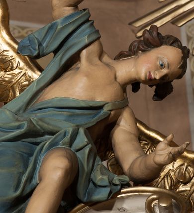 Zdjęcie nr 2: Rzeźby ścięte z tyłu, drążone, ukazane jako siedzące na przerwanym przyczółku, skierowane w stronę ołtarza. Lewa figura pochylona, z uniesioną nad głową prawą ręką i wyciągniętą przed siebie lewą. Figura anioła po prawej, lewą rękę kładzie na piersi, prawą wyciąga przed siebie. Anioły mają pociągłe twarze, z wystającymi brodami, dużymi oczami, zadartymi nosami i pełnymi ustami, okolone silnie kręconymi włosami opadającymi na kark. Anioł po lewej stronie ubrany jest w niebieską, silnie drapowaną szatę, odsłaniającą częściowo tors, przerzuconą przez prawe ramię, podtrzymywaną na lewym ramieniu wstążką, otaczającą postać w partii bioder. Anioł po prawej ubrany jest w czerwoną, ekspresyjnie fałdowaną szatę odsłaniającą tors, przerzuconą przez lewe ramię i otaczającą postać w partii bioder. Skrzydła obu aniołów rozpostarte na boki, pozłocone. Polichromia ciała naturalistyczna. 