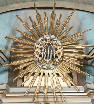 Zdjęcie nr 16: Nastawa architektoniczna, jednoosiowa, jednokondygnacyjna, ustawiona na wysokim cokole. Ołtarz w formie prostopadłościanu o ściętych z przodu narożach z prostokątną, drewnianą mensą, dekorowany trzema płycinami, na froncie monogram Marii ujęty winnym gronem. Na ołtarzu tabernakulum w formie prostopadłościennej skrzynki o ściętych przednich narożach, flankowanej parą pilastrów podtrzymujących gzyms. Drzwiczki w formie stojącego prostokąta zamkniętego półkoliście, ujęte profilowaną ramą, w polu płaskorzeźbiony kielich z hostią. 
Nastawa flankowana parą zwróconych na zewnątrz zdwojonych pilastrów i stojącą przed nimi parą kolumn o kapitelach kompozytowych. Podpory dźwigają pełne belkowanie z wyłamanym nad polem środkowym na kształt łuku odcinkowego gzymsem oraz przerwany, półkolisty przyczółek. Po zewnętrznej stronie kolumn dwie konsole, na których stoją figury św. Anny i św. Joachima. W polu głównym nisza w formie stojącego prostokąta zamkniętego łukiem półkolistym nadwieszonym z uskokiem, ujęta profilowaną ramą, dekorowana rocaille&#039;em, wewnątrz mniejszy obraz Matki Boskiej z Dzieciątkiem w kształcie stojącego prostokąta. W przyłuczach niszy dwie ukwiecone gałązki, na gzymsie uskrzydlona główka anielska, u dołu rocaille&#039;owy kartusz. W miejscu zwieńczenia, na tle okna zamontowany medalion z monogramem Marii w glorii promienistej. Struktura polichromowana w kolorze jasnoniebieskim z beżowymi detalami, ornamentyka, kolumny i profile pozłocone. 