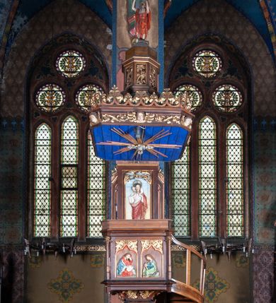 Zdjęcie nr 3: Ambona stojąca, usytuowana przy filarze z dostępem schodami z prawej strony, ustawiona na czworościennym filarze. Filar osadzony jest na szerokiej plincie i węższej bazie; trzon podzielony jest na dwie części. Poszczególne partie trzonu oddzielone są od siebie gzymsami. Naroża dolnej części artykułowane są filarkami ustawionymi na wysokich cokołach, a ścianki filarów dekorowane są neogotyckimi płycinami, zakończonymi motywem dwuliścia. Ścianki dolnej części cokołu zdobione są podwójnymi blendami. Górna część trzonu jest niższa, w narożach artykułowana pinaklami o szczytach wywiniętych do przodu. Na ściankach kwadratowe płyciny wypełnione rzeźbioną i złoconą dekoracją roślinną. Nad tą częścią trzonu osadzony jest ścięty ostrosłup czworokątny, na którym wspiera się kosz ambony. Ścianki ostrosłupa wypełnione są rzeźbionymi atrybutami ewangelistów przedstawiającymi im odpowiednio: anioła, lwa, orła i woła.

Kosz ambony założony jest na planie kwadratu, naroża flankują wąskie kolumienki wspierające niepełne belkowanie i parapet. Bazy kolumienek wysokie, sześcioboczne, wyżej zdobione rzeźbioną plecionką; trzony gładkie, kapitele zdobione motywami neogotyckimi. Na lewej i środkowej ściance kosza umieszczono po dwie prostokątne płyciny z rzeźbionymi przedstawieniami ewangelistów. Kompozycje zamknięte w profilowanych ramach w kształcie stojących prostokątów, zamkniętych oślim grzbietem i motywem akantu rozchodzącym się od niego symetrycznie na przyłucza. Ewangeliści ukazani są w pozycji siedzącej z księgami w dłoniach. Ubrani są w różnokolorowe suknie z długimi rękawami i płaszcze o złoconej bordiurze. Twarze szczupłe, okolone brodami i bujnymi oraz silnie skręconymi włosami. 

Zaplecek prostokątny, w polu w prostokątnej płycinie płaskorzeźba Chrystusa Dobrego Pasterza, u góry dekoracja złożona z astwerku i liści akantu. Baldachim na planie kwadratu o ściętych narożach. Podniebienie polichromowane na kolor ciemnoniebieski z rzeźbioną gołębicą Ducha Świętego w polu na tle promienistej glorii złożonej z wiązek promieni o nierównej długości. Fryz belkowania zdobią uskrzydlone główki aniołków, skierowane na wprost oraz dekoracja roślinna, nad belkowaniem biegnie rzeźbiony pas ażurowej koronki w kształcie wici roślinnej. Na baldachimie ośmioboczny cokół z otworami w kształcie stojących prostokątów wypełnionych ażurowym ornamentem roślinnym. Na cokole ustawiona jest ośmioboczna podstawa o uskokowej górnej części, na które stoi pełnoplastyczna figura Chrystusa Zmartwychwstałego, ubranego w czerwony płaszcz, który w lewej dłoni trzyma chorągiew, a prawą unosi do góry. Jego twarz jest podłużna, okolona zarostem, włosy długie, kręcone i bujne, a wokół głowy ma promienisty nimb. Schody z poręczą i prostymi tralkami, bez zdobień. Ambona w kolorze naturalnego drewna, ornamentyka złocona, detale srebrzone i złocone, podniebienie baldachimu polichromowane na kolor ciemnoniebieski. 
