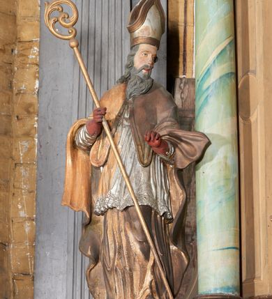 Zdjęcie nr 1: Figura drążona z tyłu; ustawiona na niskiej, nieregularnej podstawie; ukazana frontalnie, w lekkim kontrapoście, z głową zwróconą w trzech czwartych w lewo. Obie ręce ugięte, wyciągnięte przed siebie, w prawej trzyma pastorał. Twarz  pociągła, o małych opadających oczach, wąskim, niewielkim nosie i lekko rozchylonych ustach; okolona silnie falowanymi włosami i skręconą brodą opadającą na pierś, z wyraźnie zaznaczonymi długimi wąsami. Postać ubrana w silnie drapowane albę i rokietę o płaskorzeźbionym brzegu. Na ramiona ma narzuconą kapę, zapiętą pod szyją klamrą, opadającą na przedramiona i plecy, z rozwianym fragmentem na lewej ręce. Na dłoniach ma rękawiczki, na głowie infułę, z grubą lamówką, zakończoną krzyżem, na stopach ma buty. Strój niemal w całości złocony, miejscami z przetartymi srebrzeniami; rękawiczki i buty malowane w kolorze czerwonym; polichromia twarzy i włosów naturalistyczna. 






