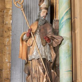 Zdjęcie nr 1: Figura drążona z tyłu; ustawiona na niskiej, nieregularnej podstawie; ukazana frontalnie, w lekkim kontrapoście, z głową zwróconą w trzech czwartych w lewo. Obie ręce ugięte, wyciągnięte przed siebie, w prawej trzyma pastorał. Twarz  pociągła, o małych opadających oczach, wąskim, niewielkim nosie i lekko rozchylonych ustach; okolona silnie falowanymi włosami i skręconą brodą opadającą na pierś, z wyraźnie zaznaczonymi długimi wąsami. Postać ubrana w silnie drapowane albę i rokietę o płaskorzeźbionym brzegu. Na ramiona ma narzuconą kapę, zapiętą pod szyją klamrą, opadającą na przedramiona i plecy, z rozwianym fragmentem na lewej ręce. Na dłoniach ma rękawiczki, na głowie infułę, z grubą lamówką, zakończoną krzyżem, na stopach ma buty. Strój niemal w całości złocony, miejscami z przetartymi srebrzeniami; rękawiczki i buty malowane w kolorze czerwonym; polichromia twarzy i włosów naturalistyczna. 






