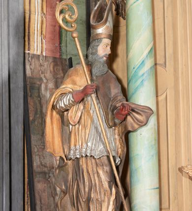 Zdjęcie nr 2: Figura drążona z tyłu; ustawiona na niskiej, nieregularnej podstawie; ukazana frontalnie, w lekkim kontrapoście, z głową zwróconą w trzech czwartych w lewo. Obie ręce ugięte, wyciągnięte przed siebie, w prawej trzyma pastorał. Twarz  pociągła, o małych opadających oczach, wąskim, niewielkim nosie i lekko rozchylonych ustach; okolona silnie falowanymi włosami i skręconą brodą opadającą na pierś, z wyraźnie zaznaczonymi długimi wąsami. Postać ubrana w silnie drapowane albę i rokietę o płaskorzeźbionym brzegu. Na ramiona ma narzuconą kapę, zapiętą pod szyją klamrą, opadającą na przedramiona i plecy, z rozwianym fragmentem na lewej ręce. Na dłoniach ma rękawiczki, na głowie infułę, z grubą lamówką, zakończoną krzyżem, na stopach ma buty. Strój niemal w całości złocony, miejscami z przetartymi srebrzeniami; rękawiczki i buty malowane w kolorze czerwonym; polichromia twarzy i włosów naturalistyczna. 






