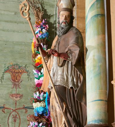 Zdjęcie nr 3: Figura drążona z tyłu; ustawiona na niskiej, nieregularnej podstawie; ukazana frontalnie, w lekkim kontrapoście, z głową zwróconą w trzech czwartych w lewo. Obie ręce ugięte, wyciągnięte przed siebie, w prawej trzyma pastorał. Twarz  pociągła, o małych opadających oczach, wąskim, niewielkim nosie i lekko rozchylonych ustach; okolona silnie falowanymi włosami i skręconą brodą opadającą na pierś, z wyraźnie zaznaczonymi długimi wąsami. Postać ubrana w silnie drapowane albę i rokietę o płaskorzeźbionym brzegu. Na ramiona ma narzuconą kapę, zapiętą pod szyją klamrą, opadającą na przedramiona i plecy, z rozwianym fragmentem na lewej ręce. Na dłoniach ma rękawiczki, na głowie infułę, z grubą lamówką, zakończoną krzyżem, na stopach ma buty. Strój niemal w całości złocony, miejscami z przetartymi srebrzeniami; rękawiczki i buty malowane w kolorze czerwonym; polichromia twarzy i włosów naturalistyczna. 






