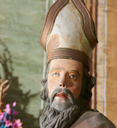 Zdjęcie nr 5: Figura drążona z tyłu; ustawiona na niskiej, nieregularnej podstawie; ukazana frontalnie, w lekkim kontrapoście, z głową zwróconą w trzech czwartych w lewo. Obie ręce ugięte, wyciągnięte przed siebie, w prawej trzyma pastorał. Twarz  pociągła, o małych opadających oczach, wąskim, niewielkim nosie i lekko rozchylonych ustach; okolona silnie falowanymi włosami i skręconą brodą opadającą na pierś, z wyraźnie zaznaczonymi długimi wąsami. Postać ubrana w silnie drapowane albę i rokietę o płaskorzeźbionym brzegu. Na ramiona ma narzuconą kapę, zapiętą pod szyją klamrą, opadającą na przedramiona i plecy, z rozwianym fragmentem na lewej ręce. Na dłoniach ma rękawiczki, na głowie infułę, z grubą lamówką, zakończoną krzyżem, na stopach ma buty. Strój niemal w całości złocony, miejscami z przetartymi srebrzeniami; rękawiczki i buty malowane w kolorze czerwonym; polichromia twarzy i włosów naturalistyczna. 






