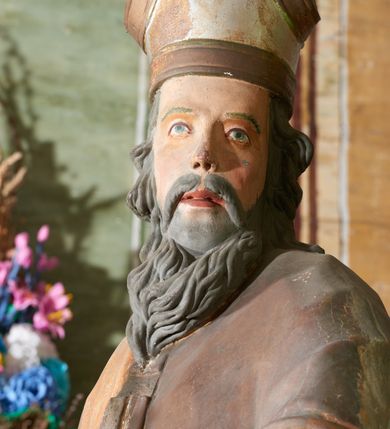 Zdjęcie nr 6: Figura drążona z tyłu; ustawiona na niskiej, nieregularnej podstawie; ukazana frontalnie, w lekkim kontrapoście, z głową zwróconą w trzech czwartych w lewo. Obie ręce ugięte, wyciągnięte przed siebie, w prawej trzyma pastorał. Twarz  pociągła, o małych opadających oczach, wąskim, niewielkim nosie i lekko rozchylonych ustach; okolona silnie falowanymi włosami i skręconą brodą opadającą na pierś, z wyraźnie zaznaczonymi długimi wąsami. Postać ubrana w silnie drapowane albę i rokietę o płaskorzeźbionym brzegu. Na ramiona ma narzuconą kapę, zapiętą pod szyją klamrą, opadającą na przedramiona i plecy, z rozwianym fragmentem na lewej ręce. Na dłoniach ma rękawiczki, na głowie infułę, z grubą lamówką, zakończoną krzyżem, na stopach ma buty. Strój niemal w całości złocony, miejscami z przetartymi srebrzeniami; rękawiczki i buty malowane w kolorze czerwonym; polichromia twarzy i włosów naturalistyczna. 






