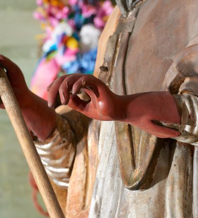 Zdjęcie nr 8: Figura drążona z tyłu; ustawiona na niskiej, nieregularnej podstawie; ukazana frontalnie, w lekkim kontrapoście, z głową zwróconą w trzech czwartych w lewo. Obie ręce ugięte, wyciągnięte przed siebie, w prawej trzyma pastorał. Twarz  pociągła, o małych opadających oczach, wąskim, niewielkim nosie i lekko rozchylonych ustach; okolona silnie falowanymi włosami i skręconą brodą opadającą na pierś, z wyraźnie zaznaczonymi długimi wąsami. Postać ubrana w silnie drapowane albę i rokietę o płaskorzeźbionym brzegu. Na ramiona ma narzuconą kapę, zapiętą pod szyją klamrą, opadającą na przedramiona i plecy, z rozwianym fragmentem na lewej ręce. Na dłoniach ma rękawiczki, na głowie infułę, z grubą lamówką, zakończoną krzyżem, na stopach ma buty. Strój niemal w całości złocony, miejscami z przetartymi srebrzeniami; rękawiczki i buty malowane w kolorze czerwonym; polichromia twarzy i włosów naturalistyczna. 






