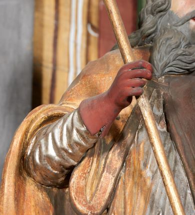 Zdjęcie nr 9: Figura drążona z tyłu; ustawiona na niskiej, nieregularnej podstawie; ukazana frontalnie, w lekkim kontrapoście, z głową zwróconą w trzech czwartych w lewo. Obie ręce ugięte, wyciągnięte przed siebie, w prawej trzyma pastorał. Twarz  pociągła, o małych opadających oczach, wąskim, niewielkim nosie i lekko rozchylonych ustach; okolona silnie falowanymi włosami i skręconą brodą opadającą na pierś, z wyraźnie zaznaczonymi długimi wąsami. Postać ubrana w silnie drapowane albę i rokietę o płaskorzeźbionym brzegu. Na ramiona ma narzuconą kapę, zapiętą pod szyją klamrą, opadającą na przedramiona i plecy, z rozwianym fragmentem na lewej ręce. Na dłoniach ma rękawiczki, na głowie infułę, z grubą lamówką, zakończoną krzyżem, na stopach ma buty. Strój niemal w całości złocony, miejscami z przetartymi srebrzeniami; rękawiczki i buty malowane w kolorze czerwonym; polichromia twarzy i włosów naturalistyczna. 






