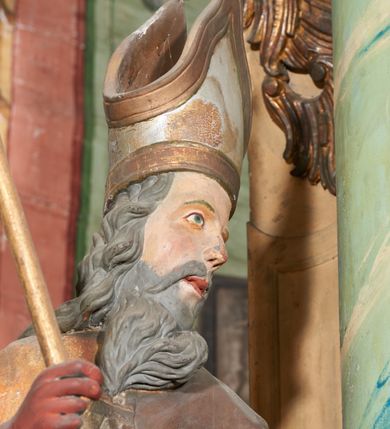 Zdjęcie nr 7: Figura drążona z tyłu; ustawiona na niskiej, nieregularnej podstawie; ukazana frontalnie, w lekkim kontrapoście, z głową zwróconą w trzech czwartych w lewo. Obie ręce ugięte, wyciągnięte przed siebie, w prawej trzyma pastorał. Twarz  pociągła, o małych opadających oczach, wąskim, niewielkim nosie i lekko rozchylonych ustach; okolona silnie falowanymi włosami i skręconą brodą opadającą na pierś, z wyraźnie zaznaczonymi długimi wąsami. Postać ubrana w silnie drapowane albę i rokietę o płaskorzeźbionym brzegu. Na ramiona ma narzuconą kapę, zapiętą pod szyją klamrą, opadającą na przedramiona i plecy, z rozwianym fragmentem na lewej ręce. Na dłoniach ma rękawiczki, na głowie infułę, z grubą lamówką, zakończoną krzyżem, na stopach ma buty. Strój niemal w całości złocony, miejscami z przetartymi srebrzeniami; rękawiczki i buty malowane w kolorze czerwonym; polichromia twarzy i włosów naturalistyczna. 






