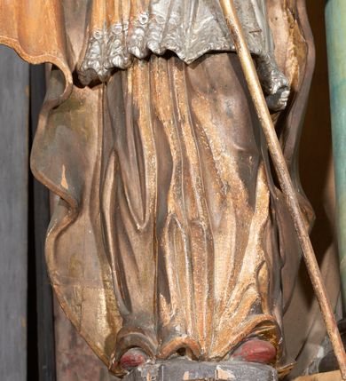 Zdjęcie nr 10: Figura drążona z tyłu; ustawiona na niskiej, nieregularnej podstawie; ukazana frontalnie, w lekkim kontrapoście, z głową zwróconą w trzech czwartych w lewo. Obie ręce ugięte, wyciągnięte przed siebie, w prawej trzyma pastorał. Twarz  pociągła, o małych opadających oczach, wąskim, niewielkim nosie i lekko rozchylonych ustach; okolona silnie falowanymi włosami i skręconą brodą opadającą na pierś, z wyraźnie zaznaczonymi długimi wąsami. Postać ubrana w silnie drapowane albę i rokietę o płaskorzeźbionym brzegu. Na ramiona ma narzuconą kapę, zapiętą pod szyją klamrą, opadającą na przedramiona i plecy, z rozwianym fragmentem na lewej ręce. Na dłoniach ma rękawiczki, na głowie infułę, z grubą lamówką, zakończoną krzyżem, na stopach ma buty. Strój niemal w całości złocony, miejscami z przetartymi srebrzeniami; rękawiczki i buty malowane w kolorze czerwonym; polichromia twarzy i włosów naturalistyczna. 






