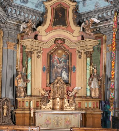 Zdjęcie nr 4: Figura drążona z tyłu; ustawiona na niskiej, nieregularnej podstawie; ukazana frontalnie, w lekkim kontrapoście, z głową zwróconą w trzech czwartych w lewo. Obie ręce ugięte, wyciągnięte przed siebie, w prawej trzyma pastorał. Twarz  pociągła, o małych opadających oczach, wąskim, niewielkim nosie i lekko rozchylonych ustach; okolona silnie falowanymi włosami i skręconą brodą opadającą na pierś, z wyraźnie zaznaczonymi długimi wąsami. Postać ubrana w silnie drapowane albę i rokietę o płaskorzeźbionym brzegu. Na ramiona ma narzuconą kapę, zapiętą pod szyją klamrą, opadającą na przedramiona i plecy, z rozwianym fragmentem na lewej ręce. Na dłoniach ma rękawiczki, na głowie infułę, z grubą lamówką, zakończoną krzyżem, na stopach ma buty. Strój niemal w całości złocony, miejscami z przetartymi srebrzeniami; rękawiczki i buty malowane w kolorze czerwonym; polichromia twarzy i włosów naturalistyczna. 






