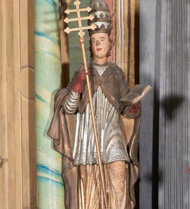 Zdjęcie nr 1: Figura drążona z tyłu; ustawiona na niskiej, nieregularnej podstawie; ukazana frontalnie, w lekkim kontrapoście, z głową zwróconą w trzech czwartych w prawo. Obie ręce ugięte, lewą podtrzymuje otwartą księgę, w prawej trzyma krzyż patriarchalny. Twarz pociągła, o małych opadających oczach, wąskim, niewielkim nosie i lekko rozchylonych ustach; okolona silnie falowanymi, krótkimi włosami. Postać ubrana w silnie drapowane albę i rokietę o płaskorzeźbionym brzegu. Na ramiona ma narzuconą kapę, zapiętą pod szyją klamrą, dekorowaną wzdłuż brzegu bordiurą, opadającą na przedramiona i plecy. Na dłoniach ma rękawiczki, na głowie potrójną tiarę, a na stopach ma buty. Strój niemal w całości złocony, miejscami z przetartymi srebrzeniami; rękawiczki i buty malowane w kolorze czerwonym; polichromia twarzy i włosów naturalistyczna. 


