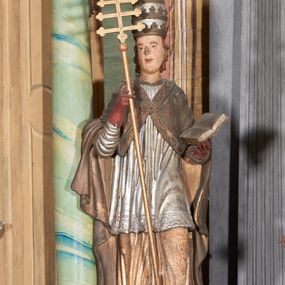 Zdjęcie nr 1: Figura drążona z tyłu; ustawiona na niskiej, nieregularnej podstawie; ukazana frontalnie, w lekkim kontrapoście, z głową zwróconą w trzech czwartych w prawo. Obie ręce ugięte, lewą podtrzymuje otwartą księgę, w prawej trzyma krzyż patriarchalny. Twarz pociągła, o małych opadających oczach, wąskim, niewielkim nosie i lekko rozchylonych ustach; okolona silnie falowanymi, krótkimi włosami. Postać ubrana w silnie drapowane albę i rokietę o płaskorzeźbionym brzegu. Na ramiona ma narzuconą kapę, zapiętą pod szyją klamrą, dekorowaną wzdłuż brzegu bordiurą, opadającą na przedramiona i plecy. Na dłoniach ma rękawiczki, na głowie potrójną tiarę, a na stopach ma buty. Strój niemal w całości złocony, miejscami z przetartymi srebrzeniami; rękawiczki i buty malowane w kolorze czerwonym; polichromia twarzy i włosów naturalistyczna. 


