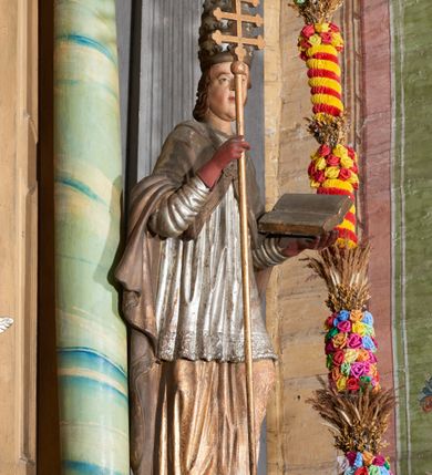 Zdjęcie nr 2: Figura drążona z tyłu; ustawiona na niskiej, nieregularnej podstawie; ukazana frontalnie, w lekkim kontrapoście, z głową zwróconą w trzech czwartych w prawo. Obie ręce ugięte, lewą podtrzymuje otwartą księgę, w prawej trzyma krzyż patriarchalny. Twarz pociągła, o małych opadających oczach, wąskim, niewielkim nosie i lekko rozchylonych ustach; okolona silnie falowanymi, krótkimi włosami. Postać ubrana w silnie drapowane albę i rokietę o płaskorzeźbionym brzegu. Na ramiona ma narzuconą kapę, zapiętą pod szyją klamrą, dekorowaną wzdłuż brzegu bordiurą, opadającą na przedramiona i plecy. Na dłoniach ma rękawiczki, na głowie potrójną tiarę, a na stopach ma buty. Strój niemal w całości złocony, miejscami z przetartymi srebrzeniami; rękawiczki i buty malowane w kolorze czerwonym; polichromia twarzy i włosów naturalistyczna. 


