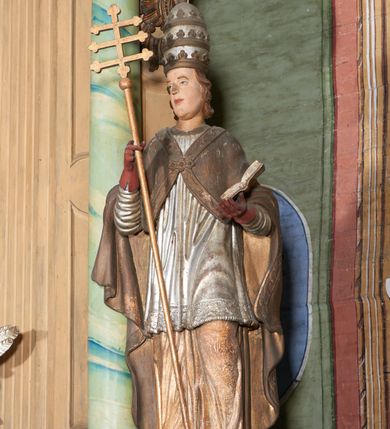 Zdjęcie nr 4: Figura drążona z tyłu; ustawiona na niskiej, nieregularnej podstawie; ukazana frontalnie, w lekkim kontrapoście, z głową zwróconą w trzech czwartych w prawo. Obie ręce ugięte, lewą podtrzymuje otwartą księgę, w prawej trzyma krzyż patriarchalny. Twarz pociągła, o małych opadających oczach, wąskim, niewielkim nosie i lekko rozchylonych ustach; okolona silnie falowanymi, krótkimi włosami. Postać ubrana w silnie drapowane albę i rokietę o płaskorzeźbionym brzegu. Na ramiona ma narzuconą kapę, zapiętą pod szyją klamrą, dekorowaną wzdłuż brzegu bordiurą, opadającą na przedramiona i plecy. Na dłoniach ma rękawiczki, na głowie potrójną tiarę, a na stopach ma buty. Strój niemal w całości złocony, miejscami z przetartymi srebrzeniami; rękawiczki i buty malowane w kolorze czerwonym; polichromia twarzy i włosów naturalistyczna. 


