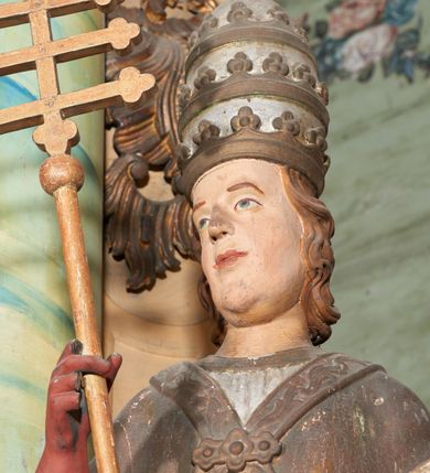 Zdjęcie nr 5: Figura drążona z tyłu; ustawiona na niskiej, nieregularnej podstawie; ukazana frontalnie, w lekkim kontrapoście, z głową zwróconą w trzech czwartych w prawo. Obie ręce ugięte, lewą podtrzymuje otwartą księgę, w prawej trzyma krzyż patriarchalny. Twarz pociągła, o małych opadających oczach, wąskim, niewielkim nosie i lekko rozchylonych ustach; okolona silnie falowanymi, krótkimi włosami. Postać ubrana w silnie drapowane albę i rokietę o płaskorzeźbionym brzegu. Na ramiona ma narzuconą kapę, zapiętą pod szyją klamrą, dekorowaną wzdłuż brzegu bordiurą, opadającą na przedramiona i plecy. Na dłoniach ma rękawiczki, na głowie potrójną tiarę, a na stopach ma buty. Strój niemal w całości złocony, miejscami z przetartymi srebrzeniami; rękawiczki i buty malowane w kolorze czerwonym; polichromia twarzy i włosów naturalistyczna. 


