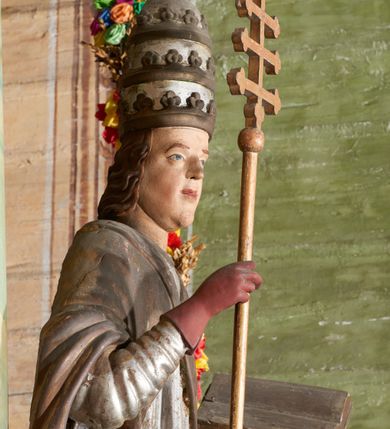 Zdjęcie nr 6: Figura drążona z tyłu; ustawiona na niskiej, nieregularnej podstawie; ukazana frontalnie, w lekkim kontrapoście, z głową zwróconą w trzech czwartych w prawo. Obie ręce ugięte, lewą podtrzymuje otwartą księgę, w prawej trzyma krzyż patriarchalny. Twarz pociągła, o małych opadających oczach, wąskim, niewielkim nosie i lekko rozchylonych ustach; okolona silnie falowanymi, krótkimi włosami. Postać ubrana w silnie drapowane albę i rokietę o płaskorzeźbionym brzegu. Na ramiona ma narzuconą kapę, zapiętą pod szyją klamrą, dekorowaną wzdłuż brzegu bordiurą, opadającą na przedramiona i plecy. Na dłoniach ma rękawiczki, na głowie potrójną tiarę, a na stopach ma buty. Strój niemal w całości złocony, miejscami z przetartymi srebrzeniami; rękawiczki i buty malowane w kolorze czerwonym; polichromia twarzy i włosów naturalistyczna. 


