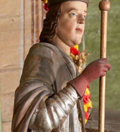 Zdjęcie nr 7: Figura drążona z tyłu; ustawiona na niskiej, nieregularnej podstawie; ukazana frontalnie, w lekkim kontrapoście, z głową zwróconą w trzech czwartych w prawo. Obie ręce ugięte, lewą podtrzymuje otwartą księgę, w prawej trzyma krzyż patriarchalny. Twarz pociągła, o małych opadających oczach, wąskim, niewielkim nosie i lekko rozchylonych ustach; okolona silnie falowanymi, krótkimi włosami. Postać ubrana w silnie drapowane albę i rokietę o płaskorzeźbionym brzegu. Na ramiona ma narzuconą kapę, zapiętą pod szyją klamrą, dekorowaną wzdłuż brzegu bordiurą, opadającą na przedramiona i plecy. Na dłoniach ma rękawiczki, na głowie potrójną tiarę, a na stopach ma buty. Strój niemal w całości złocony, miejscami z przetartymi srebrzeniami; rękawiczki i buty malowane w kolorze czerwonym; polichromia twarzy i włosów naturalistyczna. 


