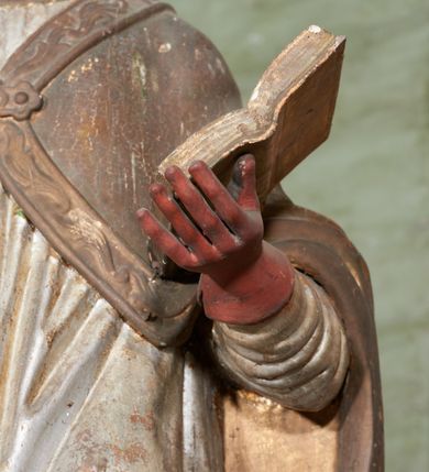 Zdjęcie nr 8: Figura drążona z tyłu; ustawiona na niskiej, nieregularnej podstawie; ukazana frontalnie, w lekkim kontrapoście, z głową zwróconą w trzech czwartych w prawo. Obie ręce ugięte, lewą podtrzymuje otwartą księgę, w prawej trzyma krzyż patriarchalny. Twarz pociągła, o małych opadających oczach, wąskim, niewielkim nosie i lekko rozchylonych ustach; okolona silnie falowanymi, krótkimi włosami. Postać ubrana w silnie drapowane albę i rokietę o płaskorzeźbionym brzegu. Na ramiona ma narzuconą kapę, zapiętą pod szyją klamrą, dekorowaną wzdłuż brzegu bordiurą, opadającą na przedramiona i plecy. Na dłoniach ma rękawiczki, na głowie potrójną tiarę, a na stopach ma buty. Strój niemal w całości złocony, miejscami z przetartymi srebrzeniami; rękawiczki i buty malowane w kolorze czerwonym; polichromia twarzy i włosów naturalistyczna. 


