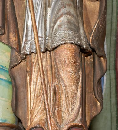 Zdjęcie nr 9: Figura drążona z tyłu; ustawiona na niskiej, nieregularnej podstawie; ukazana frontalnie, w lekkim kontrapoście, z głową zwróconą w trzech czwartych w prawo. Obie ręce ugięte, lewą podtrzymuje otwartą księgę, w prawej trzyma krzyż patriarchalny. Twarz pociągła, o małych opadających oczach, wąskim, niewielkim nosie i lekko rozchylonych ustach; okolona silnie falowanymi, krótkimi włosami. Postać ubrana w silnie drapowane albę i rokietę o płaskorzeźbionym brzegu. Na ramiona ma narzuconą kapę, zapiętą pod szyją klamrą, dekorowaną wzdłuż brzegu bordiurą, opadającą na przedramiona i plecy. Na dłoniach ma rękawiczki, na głowie potrójną tiarę, a na stopach ma buty. Strój niemal w całości złocony, miejscami z przetartymi srebrzeniami; rękawiczki i buty malowane w kolorze czerwonym; polichromia twarzy i włosów naturalistyczna. 


