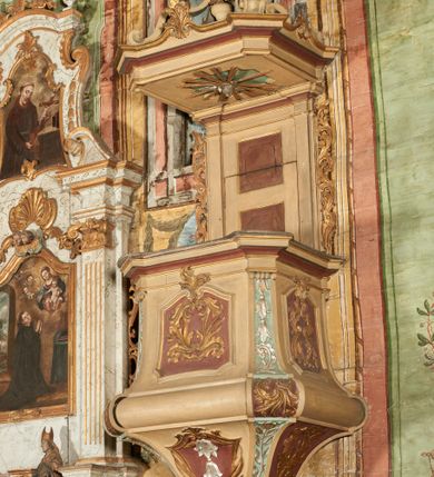 Zdjęcie nr 3: Ambona przyścienna, wisząca, z dostępem od zakrystii dzięki schodom, przez drzwiczki zamontowane w zaplecku. Kosz na planie prostokąta o ściętych narożach, rozszerzającego się ku dołowi, na podstawie w kształcie wałka, z podwieszeniem w formie zbliżonej do stożka o ściętych narożach, wklęsło-wypukłych ściankach, zakończonym szyszką przypominającą tralkę. Parapet w formie belkowania, w miejscu ściętych naroży kosza spływy wolutowe, przechodzące na wałek i podwieszenie, sięgając szyszki, dekorowane płycinami z kampanulami, rocaille'm i zwisami roślinnymi. Ścianki kosza zdobione płycinami w formie kwadratów, o ściętych dolnych narożach, zamkniętych stykającymi się falistymi wolutami z dekoracją roślinną; w polach wypełnione rocaille'm i wiciami roślinnymi. Podwieszenie kosza od frontu dekorowane roślinnym kartuszem z kampanulami w polu, na pozostałych ściankach rocaille'm i ornamentem roślinnym. Zaplecek wąski, ujęty pilastrami dźwigającymi pełne belkowanie. W polu dekorowany dwoma płycinami; z zamontowanymi drzwiczkami. Flankowany uszami utworzonymi z ornamentu roślinnego. Baldachim na planie prostokąta o ściętych narożach; obwiedziony pełnym belkowaniem, wyłamanym w miejscu ściętych naroży i z trzech stron zakończonym szczytami z palmetami. Na baldachimie cztery kabłąki w formie wolut i cęg, stanowiące podstawę dla gorejącego wazonu. Na podniebieniu baldachimu płycina z rzeźbioną gołębicą Ducha Świętego w glorii promienistej. Struktura ambony polichromowana w kolorze beżowym, płyciny i fryzy jasnoczerwonym, spływy wolutowe jasnoniebieskim; profile i ornamentyka złocone i srebrzone. 

