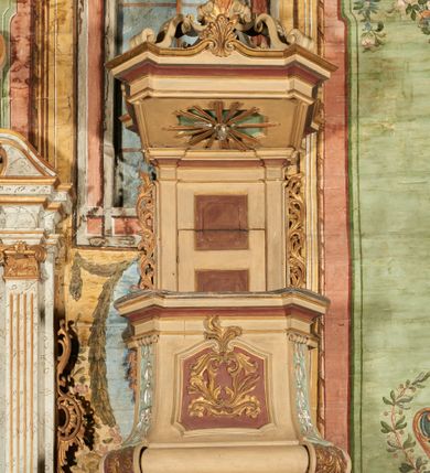 Zdjęcie nr 2: Ambona przyścienna, wisząca, z dostępem od zakrystii dzięki schodom, przez drzwiczki zamontowane w zaplecku. Kosz na planie prostokąta o ściętych narożach, rozszerzającego się ku dołowi, na podstawie w kształcie wałka, z podwieszeniem w formie zbliżonej do stożka o ściętych narożach, wklęsło-wypukłych ściankach, zakończonym szyszką przypominającą tralkę. Parapet w formie belkowania, w miejscu ściętych naroży kosza spływy wolutowe, przechodzące na wałek i podwieszenie, sięgając szyszki, dekorowane płycinami z kampanulami, rocaille&#039;m i zwisami roślinnymi. Ścianki kosza zdobione płycinami w formie kwadratów, o ściętych dolnych narożach, zamkniętych stykającymi się falistymi wolutami z dekoracją roślinną; w polach wypełnione rocaille&#039;m i wiciami roślinnymi. Podwieszenie kosza od frontu dekorowane roślinnym kartuszem z kampanulami w polu, na pozostałych ściankach rocaille&#039;m i ornamentem roślinnym. Zaplecek wąski, ujęty pilastrami dźwigającymi pełne belkowanie. W polu dekorowany dwoma płycinami; z zamontowanymi drzwiczkami. Flankowany uszami utworzonymi z ornamentu roślinnego. Baldachim na planie prostokąta o ściętych narożach; obwiedziony pełnym belkowaniem, wyłamanym w miejscu ściętych naroży i z trzech stron zakończonym szczytami z palmetami. Na baldachimie cztery kabłąki w formie wolut i cęg, stanowiące podstawę dla gorejącego wazonu. Na podniebieniu baldachimu płycina z rzeźbioną gołębicą Ducha Świętego w glorii promienistej. Struktura ambony polichromowana w kolorze beżowym, płyciny i fryzy jasnoczerwonym, spływy wolutowe jasnoniebieskim; profile i ornamentyka złocone i srebrzone. 

